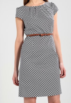 Femme portant une robe noire et blanche à motif géométrique, longueur genou, manches courtes, avec une fine ceinture marron à la taille.