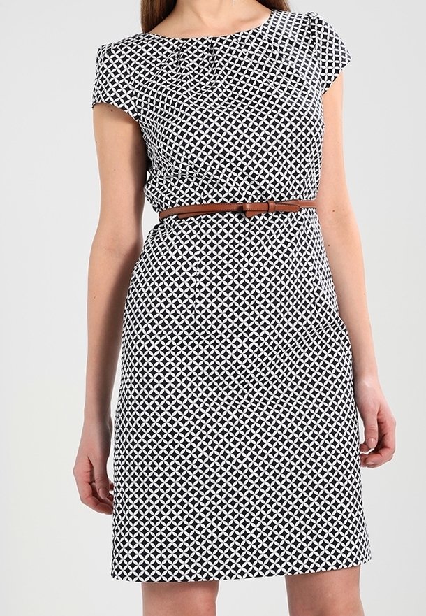 Femme portant une robe noire et blanche à motif géométrique, longueur genou, manches courtes, avec une fine ceinture marron à la taille.