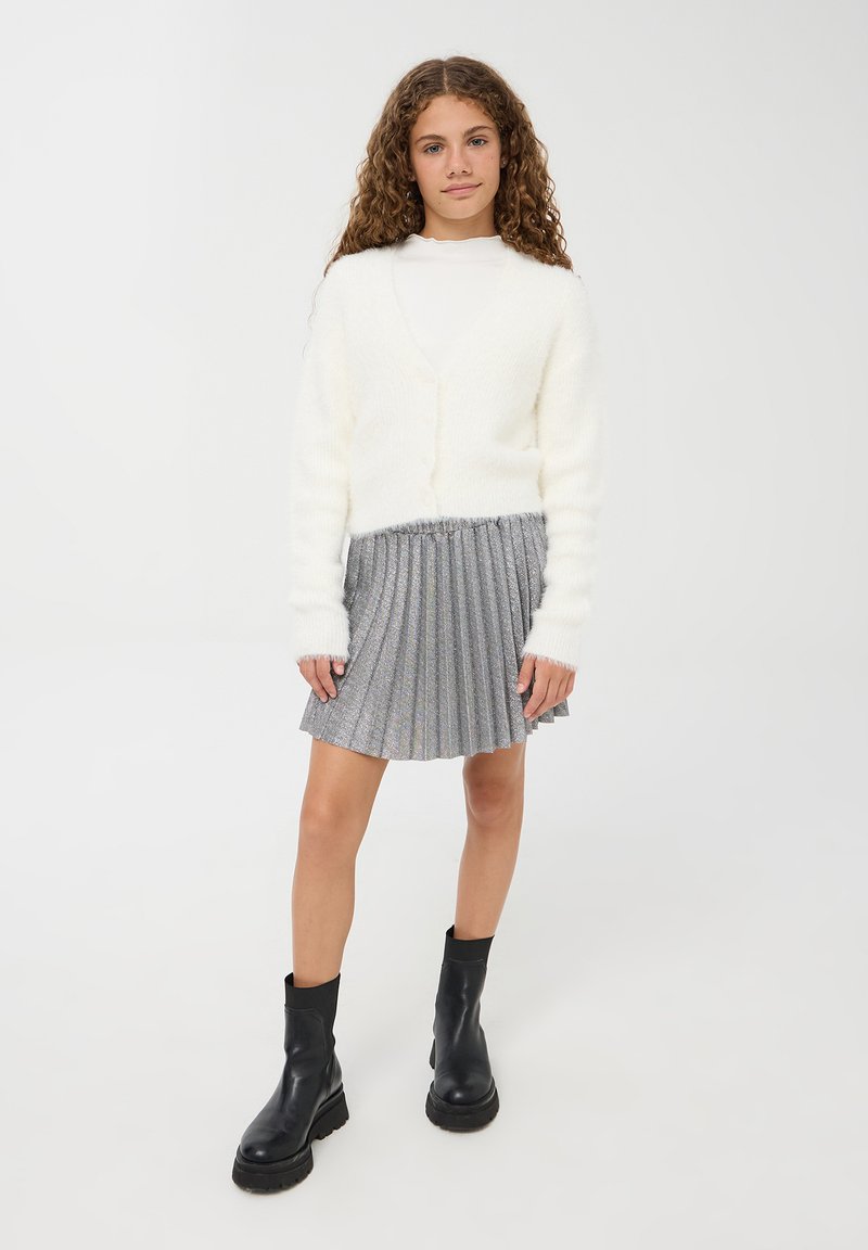 Cardigan bianco e peloso, top a maniche lunghe aderente, gonna plissettata argento e stivali neri alla caviglia con suole spesse.