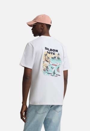 Hombre de espaldas que lleva una camiseta blanca con un gráfico colorido de la playa de la Riviera Francesa, vaqueros azul claro y una gorra rosa.
