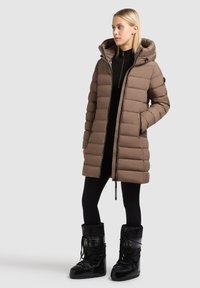 Hnedý nadýchany kabát s kapucňou, so zipsom a dlhými rukávmi; obsahuje vertikálne švy a predné vrecká; nosený s čiernymi legínami a topánkami.