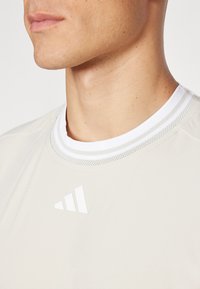 Camașă sport de culoare bej deschis, cu guler rotund, care prezintă dungi albe și gri. Include un logo Adidas alb pe partea superioară stângă. Textură netedă.