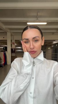 Hvit linneskjorte med høy krage og knappelukking, med lange ermer og en lett teksturert overflate. Modellen poserer med hånden mot ansiktet.