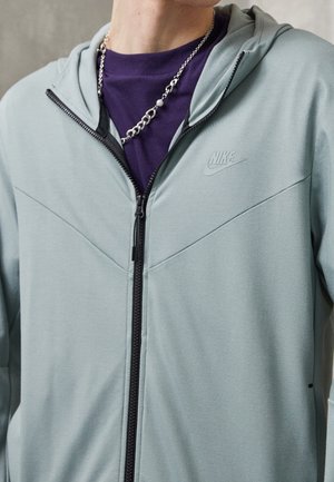 Homme portant un sweat à capuche zippé Nike gris clair sur un t-shirt violet et un collier chaîne argenté, recadré au niveau du cou et de la poitrine.