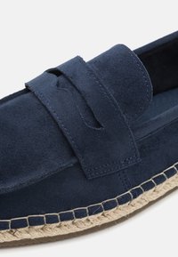 Tengerészkék velúr, slip-on loafer, oldalsó részekkel, juta kötél talppal és kontrasztos varrással az élein. Texturált felület, hétköznapi dizájn.