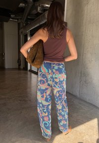 Γυναίκα που στέκεται σε ένα διάδρομο, φοράει καφέ αμάνικο τοπ, πολύχρωμο παντελόνι με σχέδια, καφέ τακούνια και κρατάει μια μεγάλη καφέ τσάντα στον ώμο της.