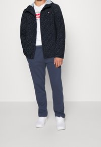 Námořnicky modrá bunda s vzorem a světle modrou kapucí, bílé tričko s potiskem a tmavě šedé tepláky. Nošeno s bílými teniskami.