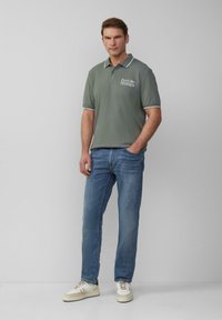 Man in grijze Ford Mustang polo, blauwe spijkerbroek en witte sneakers staat met één hand in zijn zak tegen een effen grijze achtergrond.