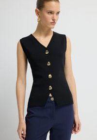Czarny bezrękawnik z dekoltem w kształcie litery V i zapięciem na guziki z przodu, z pięcioma złotymi guzikami, w połączeniu z granatowymi spodniami.
