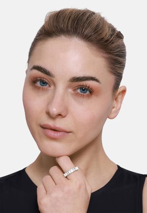 Anello in argento con gemme trasparenti rettangolari incastonate in una fascia dritta. Modella in posa con la mano sul mento, indossando un top nero.