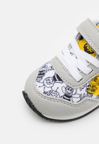 Grijze suède sneaker met een afdruk van een wit en zwart stripfiguur, witte veters en een gele accentpaneel. Rubberen zool.
