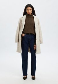 Bézs gyapjú kabát, sötétbarna garbó pulóver és sötétkék, egyenes szárú farmernadrág. Barna, magas sarkú cipő. Minimalista dizájn, sima textúrák.