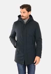 Cappotto imbottito blu scuro con cappuccio, dotato di chiusura con zip, tasche laterali e una texture liscia, indossato su un dolcevita grigio.