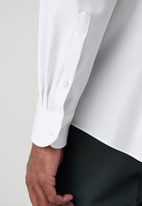 Gros plan sur la main d'une personne portant une chemise blanche à manches longues avec des poignets boutonnés et un pantalon foncé.