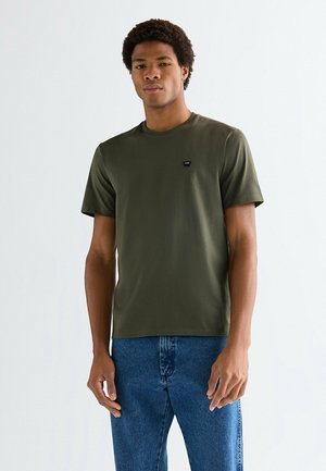 Młody mężczyzna z krótkimi kręconymi włosami, ubrany w oliwkowozieloną koszulkę i niebieskie dżinsy, stojący na jasnym, jednolitym tle.