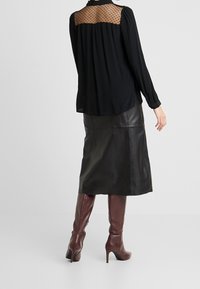 Zwarte blouse met doorschijnende stippen-yoke, gecombineerd met een zwarte leren midi-rok en bordeauxrode laarzen met hoge hakken tot de knie. Lange mouwen en een relaxte pasvorm.