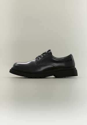 Chaussure de ville en cuir noir poli avec semelle épaisse, laçage à l'avant, et logo "REPLAY" embossé sur le côté du talon.