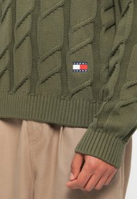 Olivgrüner, gestrickter Kabelpullover mit geripptem Saum, vorne mit einem "TOMMY JEANS"-Logopatch in Blau, Weiß und Rot.