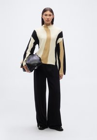 Pull en maille avec des motifs abstraits noirs, beiges et crème, associé à un pantalon large noir et tenant un sac à main noir élégant.