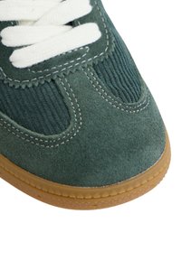 Zapatillas de ante verde con un panel de corduroy texturizado, cordones blancos y suela de goma. La parte superior presenta costuras visibles y acentos lisos.