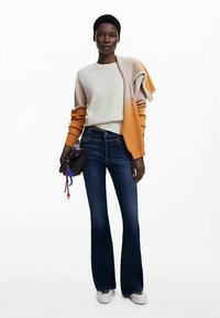 Flerfärgad tröja med orange och beige nyanser, krämfärgad skjorta, utsvängda blå jeans och vita sneakers. Håller i en liten mörk handväska med färgglada detaljer.