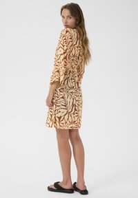 Vestido envolvente en crema con patrón en óxido, con mangas cortas, una silueta relajada y una cintura ajustada. Combinado con sandalias negras.