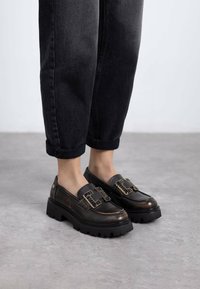 Mocasines de cuero negro con suela gruesa negra, que presentan acentos de hardware dorado y detalles texturizados, combinados con jeans negros con dobladillo.
