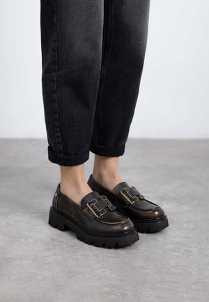 Mocasines de cuero negro con suela gruesa negra, que presentan acentos de hardware dorado y detalles texturizados, combinados con jeans negros con dobladillo.