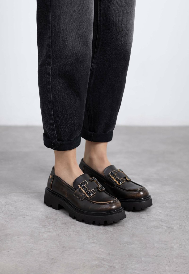 Mocasines de cuero negro con suela gruesa negra, que presentan acentos de hardware dorado y detalles texturizados, combinados con jeans negros con dobladillo.
