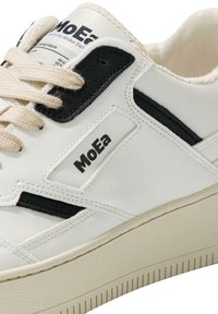 White sneaker featuring black accents, textured rubber sole, and cream laces. Branding label positioned on the side adds detail.
