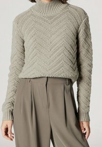 Gestrickter Rollkragenpullover in Hellgrau mit Fischgrätmuster. Verfügt über eine lockere Passform und gerippte Bündchen. Kombiniert mit beigen, plissierten Hosen.