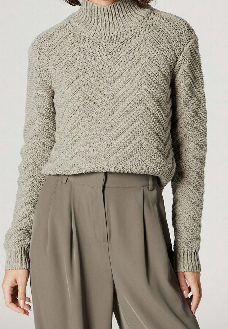 Gestrickter Rollkragenpullover in Hellgrau mit Fischgrätmuster. Verfügt über eine lockere Passform und gerippte Bündchen. Kombiniert mit beigen, plissierten Hosen.