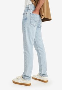 Person som bär ljusblå raka jeans och vita sneakers med gummisålar, hand i bakfickan, håller en brun skjorta över axeln.