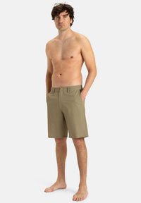 Knee-dlhé khaki šortky s gombíkovým zapínaním, klasickými vreckami a hladkou textúrou. Model stojí naboso s rukami vo vreckách.