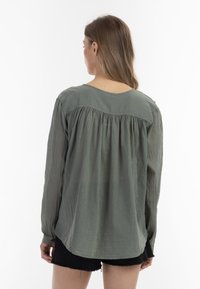 Blusa a maniche lunghe di colore verde oliva, realizzata in tessuto leggero, con una cucitura arricciata sul retro e un orlo curvo. Abbinata a pantaloni corti neri.