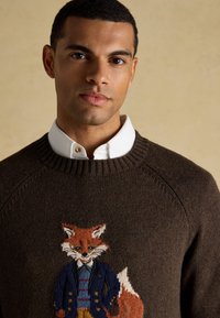 Man with short hair wearing a white collared shirt under a brown sweater featuring an embroidered fox in a blue jacket and striped shirt.