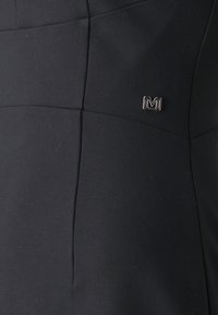 Svart kjole med en strømlinjeformet design og teksturerte paneler. En markant sølvlogo på forsiden tilfører detaljer til stoffet.