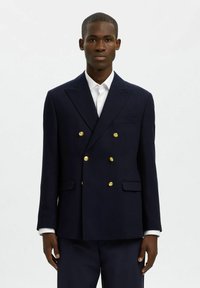 Granatowy marynarski dwurzędowy blezer z złotymi guzikami, ze zdefiniowanymi ramionami i dwoma przednimi kieszeniami. Noszony na białej koszuli.