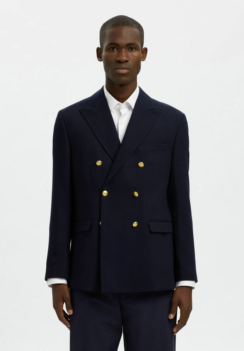 Granatowy marynarski dwurzędowy blezer z złotymi guzikami, ze zdefiniowanymi ramionami i dwoma przednimi kieszeniami. Noszony na białej koszuli.