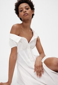 Witte off-shoulder jurk met een knoopsluiting aan de voorkant, korte mouwen en een losse pasvorm, gemaakt van gladde stof met een subtiele glans.