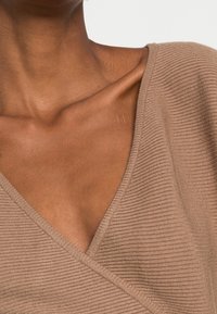 Beige overslagtop gemaakt van geribd stof; het V-halsontwerp benadrukt een gestructureerd oppervlak. Zacht en aansluitend, met schuine naden bij de halslijn.