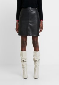 Žena v černé kožené minisukni, černém topu s dlouhým rukávem a růžovými manžetami, a volných bílých vysokých botách až ke kolenům stojí na bílém pozadí.