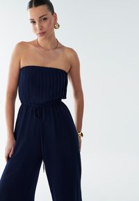 Temno modri kombinezon brez naramnic z nagubanim zgornjim delom, elastičnim pasom in vrvico za zavezovanje. Zlati okrogli uhani in gladka kovinska ogrlica.