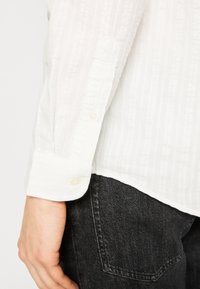 Chemise blanche en tissu boutonnée avec un motif texturé, manches à poignets et deux boutons, associée à un pantalon en denim foncé.