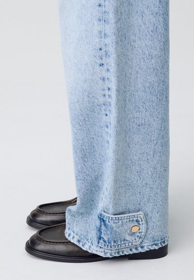 Jeans in denim blu chiaro con taglio dritto, caratterizzati da un orlo sfilacciato e dettaglio con bottone dorato, abbinati a mocassini neri.