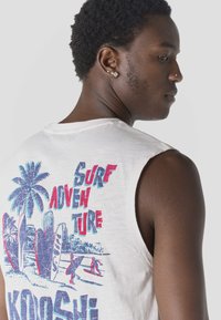 Joven con cabello corto y rizado, vistiendo una camiseta sin mangas blanca con un gráfico colorido de "Aventura de Surf" con playa y tabla de surf en la espalda.