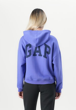 Person with long blonde hair wears a purple GAP hoodie and black pants, facing away against a plain white background.