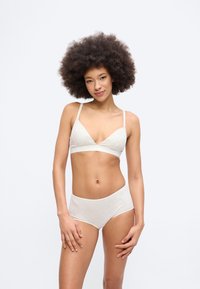 Γυναίκα με αφροαμερικανικό χτένισμα, που φοράει ανοιχτό γκρι μπράλετ και ασορτί εσώρουχα, στέκεται μπροστά σε απλό λευκό φόντο.