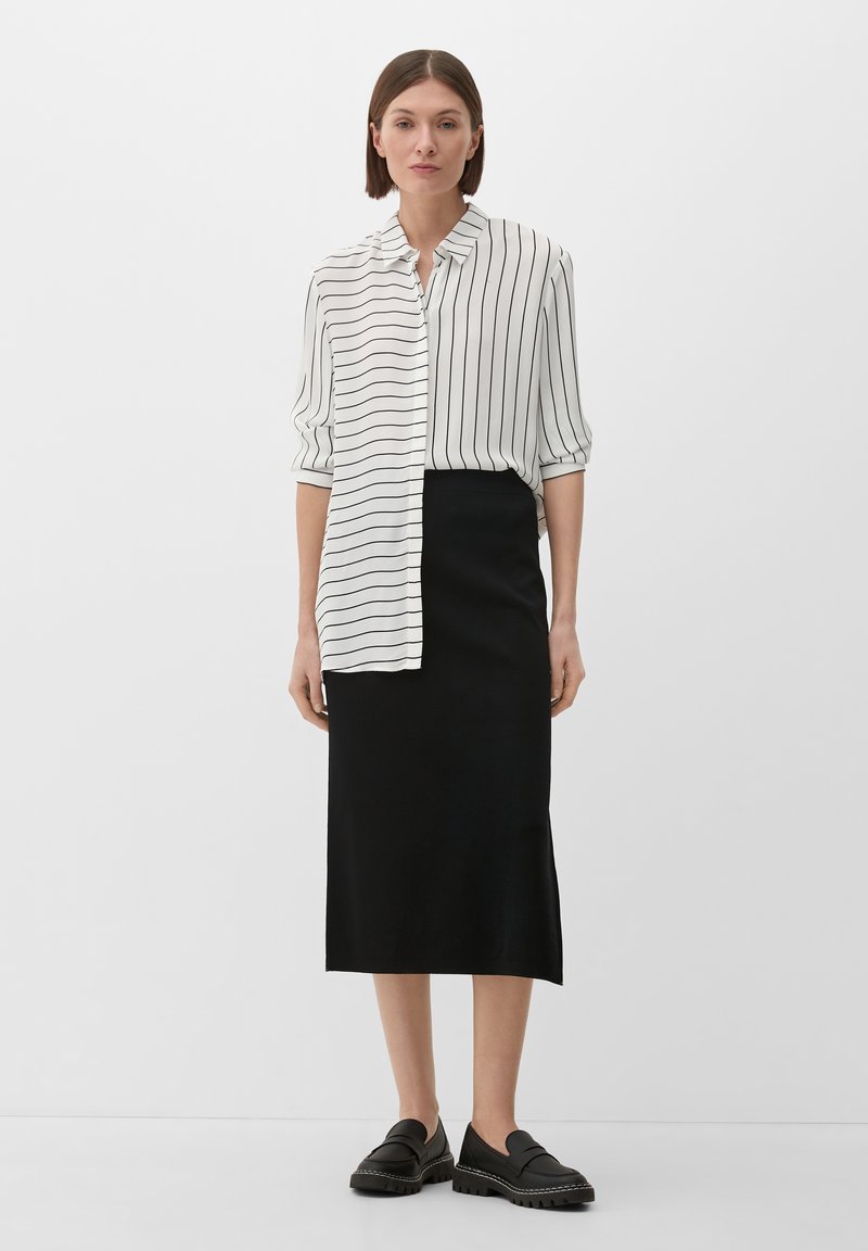 Gestreifte Bluse in Weiß und Schwarz mit Kragen, kombiniert mit einem schwarzen Midirock und schwarzen Slippers mit strukturierten Sohlen.