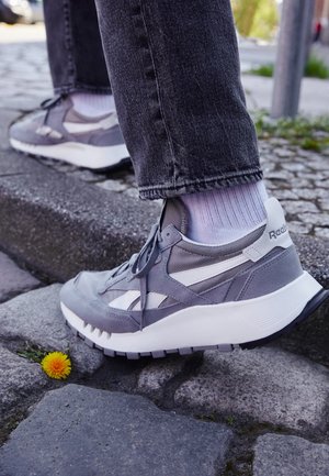 Personne portant des baskets Reebok grises et blanches marchant sur un pavé en pierre près d’une petite fleur jaune.